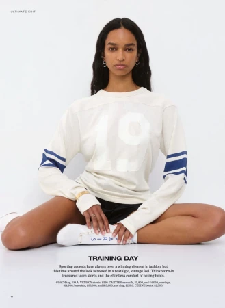 Model seated cross-legged wearing a cream sweater with blue sleeve stripes and dark shorts against a seamless white background.