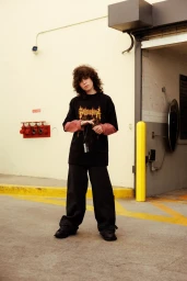 A person with curly hair wearing a black graphic t-shirt over a pink long-sleeve shirt and dark wide-leg trousers standing outdoors.