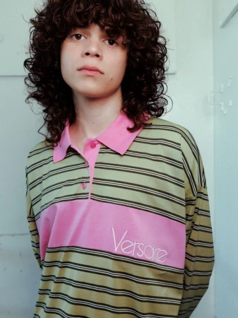 Young man with curly brown hair wearing a striped polo shirt with pink panels and embroidered text against a pale wall.