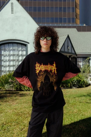 Young person with curly brown hair wearing sunglasses and black graphic t-shirt standing outside a suburban house