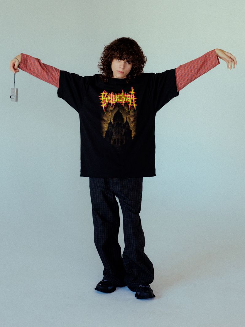 A young person with curly brown hair wearing a black t-shirt over a red long-sleeve shirt and dark trousers against a white backdrop.