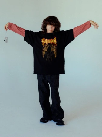 A young person with curly brown hair wearing a black t-shirt over a red long-sleeve shirt and dark trousers against a white backdrop.