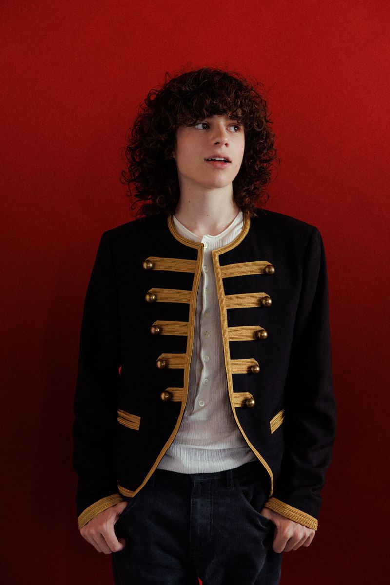 Portrait of Woody Norman with curly hair wearing a black jacket with gold trim against a red background.
