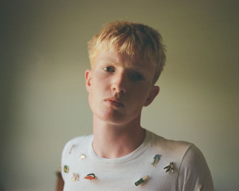 Portrait of a young man with blonde hair wearing a white t-shirt decorated with small enamel pins against a neutral wall