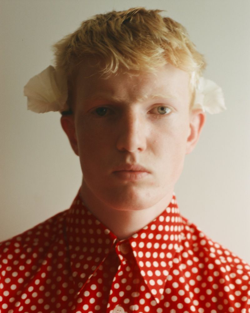 Close-up portrait of a fair-haired young man wearing a red and white polka dot shirt with white paper tucked behind his ears