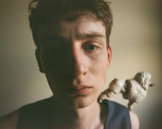 Close-up portrait of a young man wearing a dark sleeveless top with a small figurine on his shoulder against a plain background
