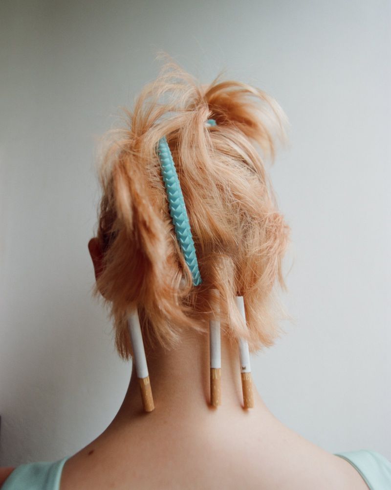 Rear view of a person with blonde hair held up by a light blue hair clip and cigarettes tucked into the hair strands
