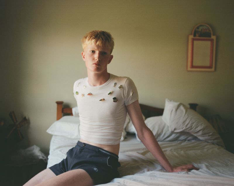 Young blonde man seated on a bed wearing a white t-shirt and dark shorts for Office Magazine