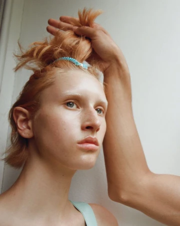 Close-up portrait of a young person with red hair tied with a blue band while a hand holds a section of hair against a white wall.