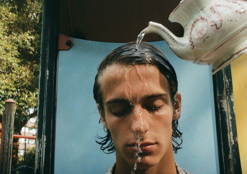Close-up of a young man having water poured onto his head from a decorative teapot against an outdoor playground structure.