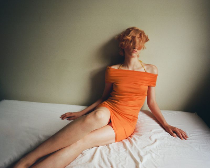 Model with orange hair wearing an orange off-the-shoulder dress seated on a bed against a neutral wall
