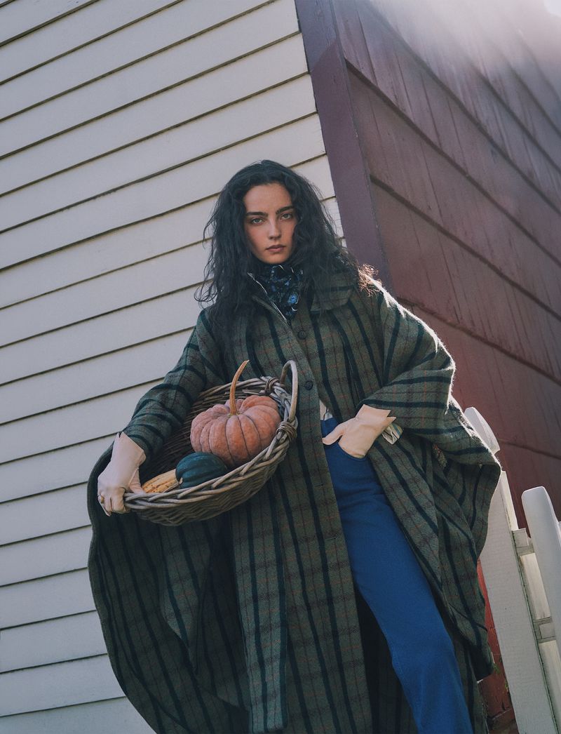 Model with long dark hair wearing a plaid green coat holding a basket of pumpkins outdoors against a building