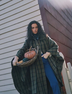 Model with long dark hair wearing a plaid green coat holding a basket of pumpkins outdoors against a building