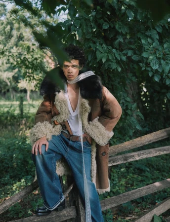 Model wearing a shearling-lined brown coat and blue jeans posing against a wooden fence surrounded by lush green foliage