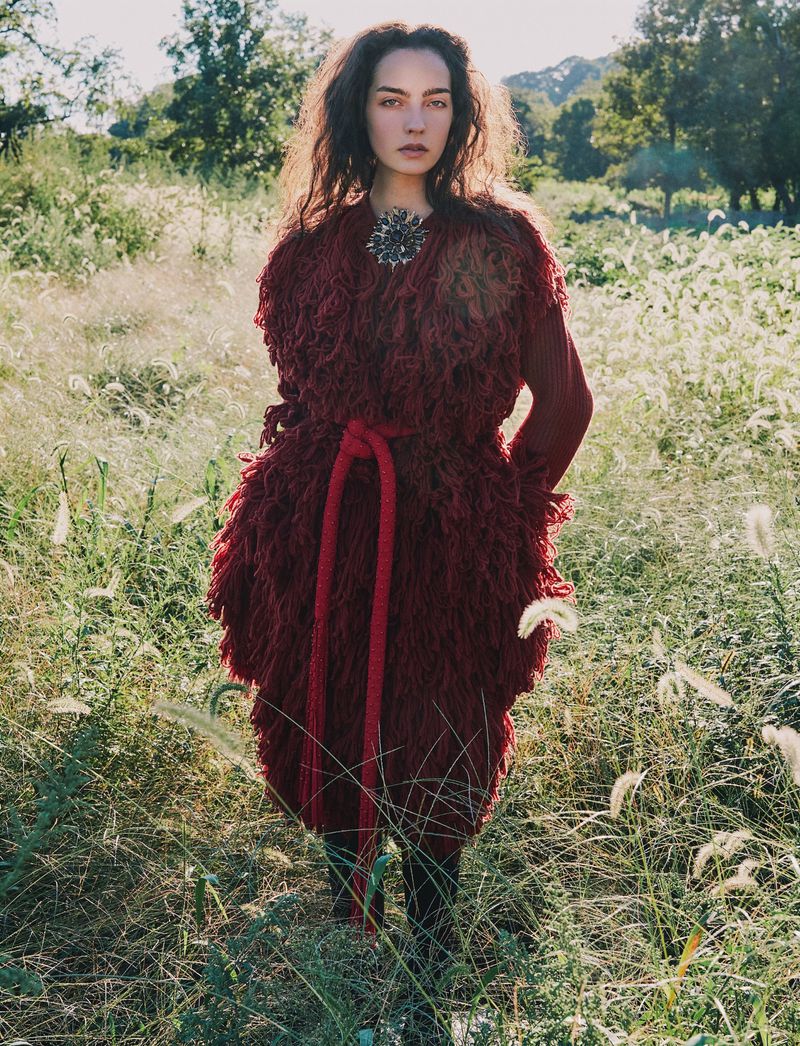 Woman standing in a tall grassy field wearing a textured burgundy fringed coat with a rope belt.