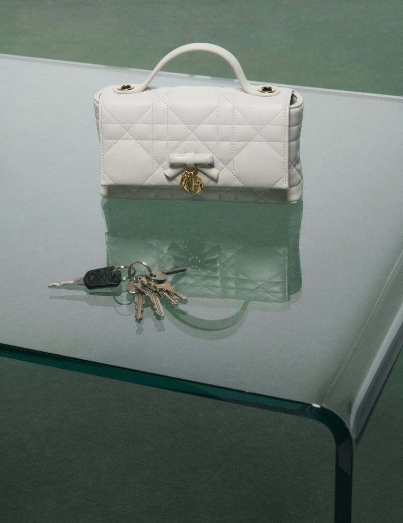White quilted handbag with a bow and gold charm resting on a glass table alongside a set of keys.