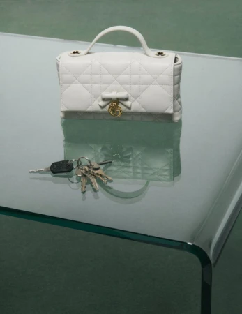 White quilted handbag with a bow and gold charm resting on a glass table alongside a set of keys.