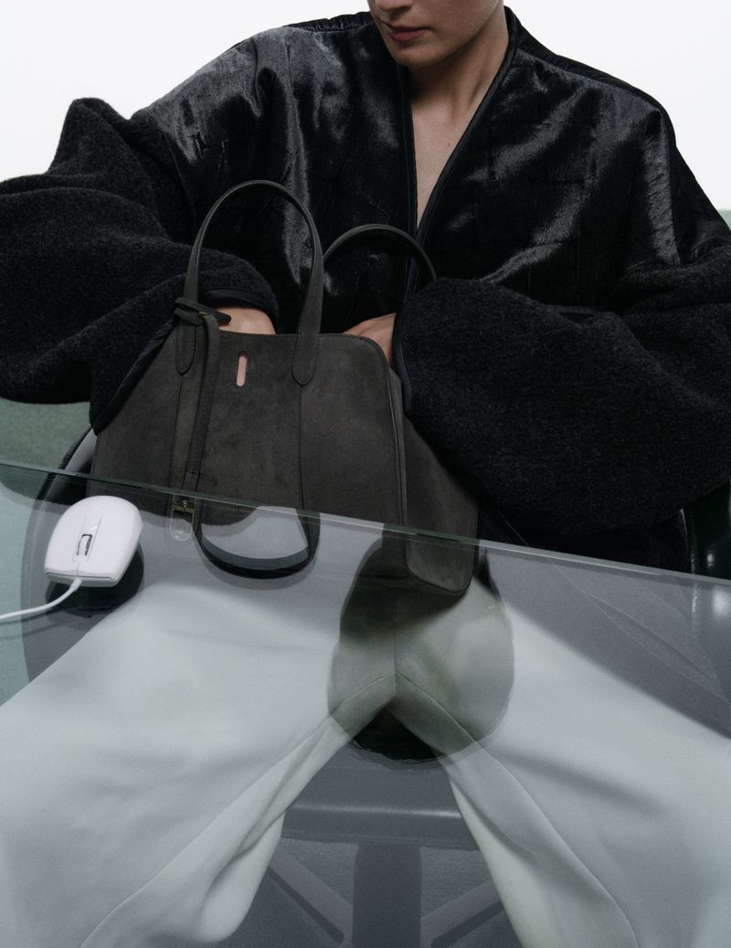 Model wearing a black jacket holding a dark handbag behind a glass desk with a white computer mouse