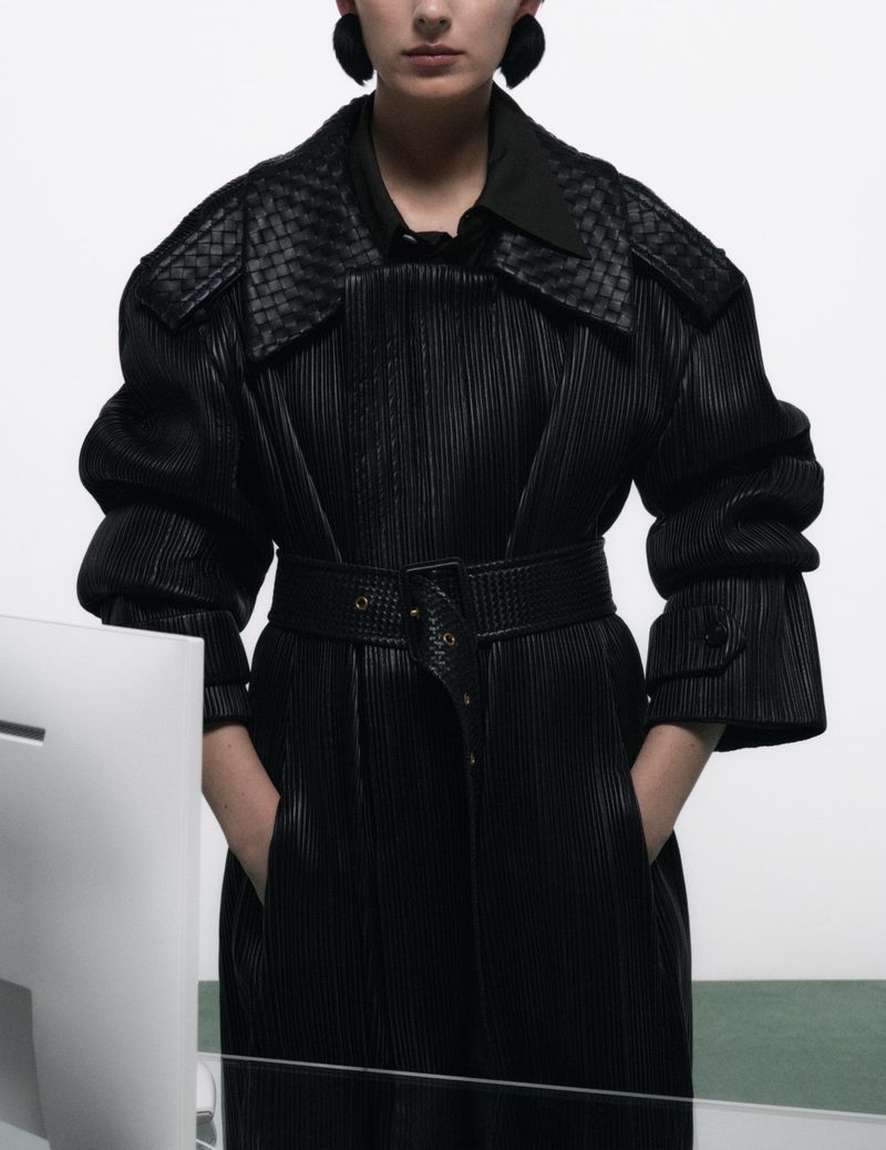 Model wearing a textured black belted coat with dark sculptural earrings against a plain background.