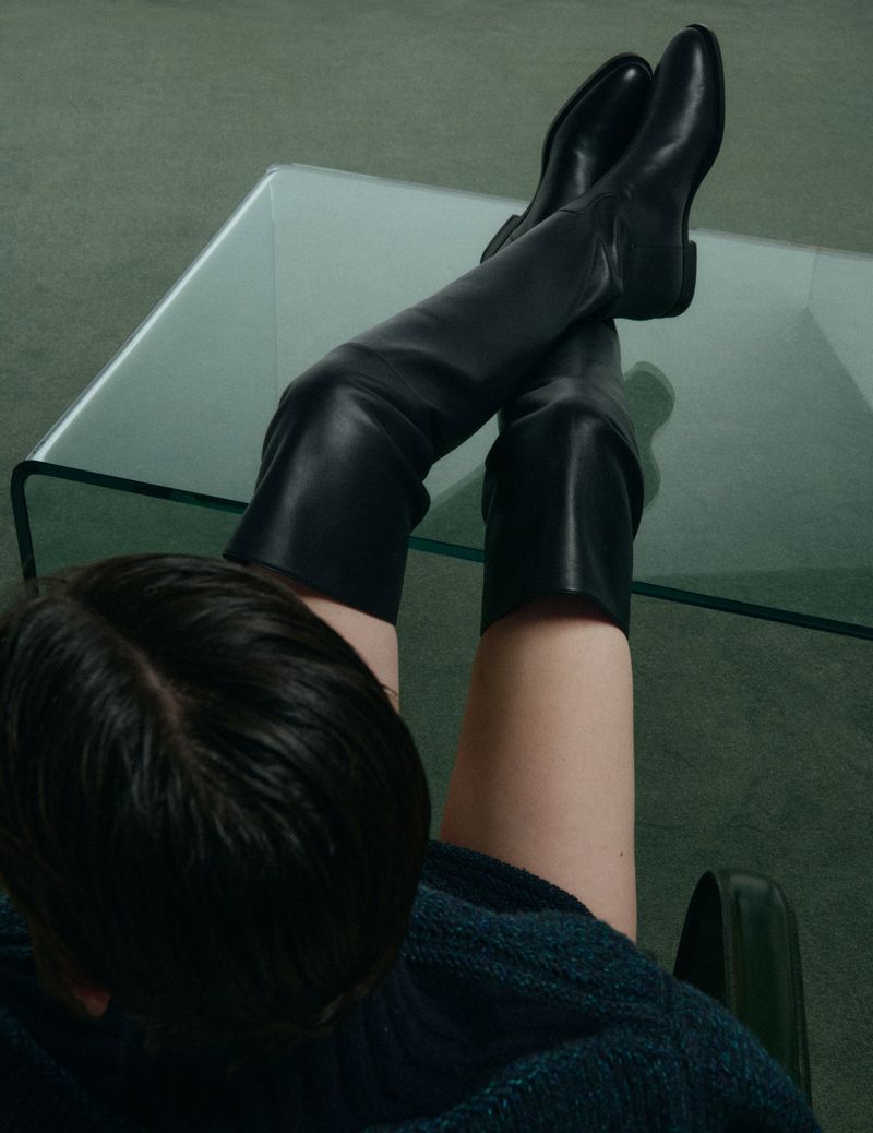 Person with dark hair wearing black knee-high leather boots resting feet on a glass table in a green carpeted room.