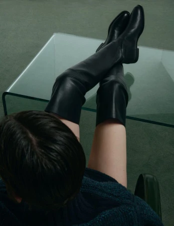 Person with dark hair wearing black knee-high leather boots resting feet on a glass table in a green carpeted room.
