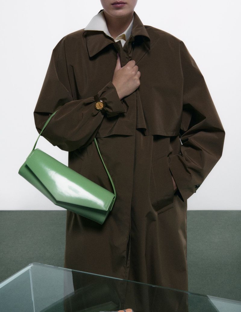 Model wearing a dark brown coat holding a light green shoulder bag against a white wall and green floor.