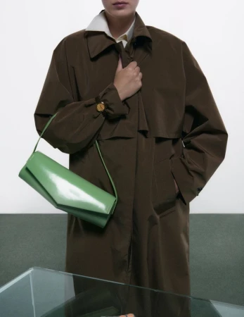 Model wearing a dark brown coat holding a light green shoulder bag against a white wall and green floor.