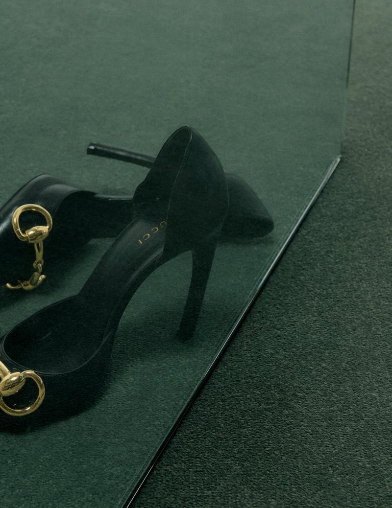 Black high heel shoes with gold horsebit hardware resting on a dark green textured carpet next to a mirror