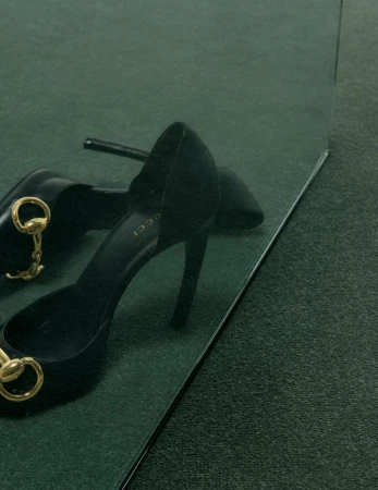 Black high heel shoes with gold horsebit hardware resting on a dark green textured carpet next to a mirror