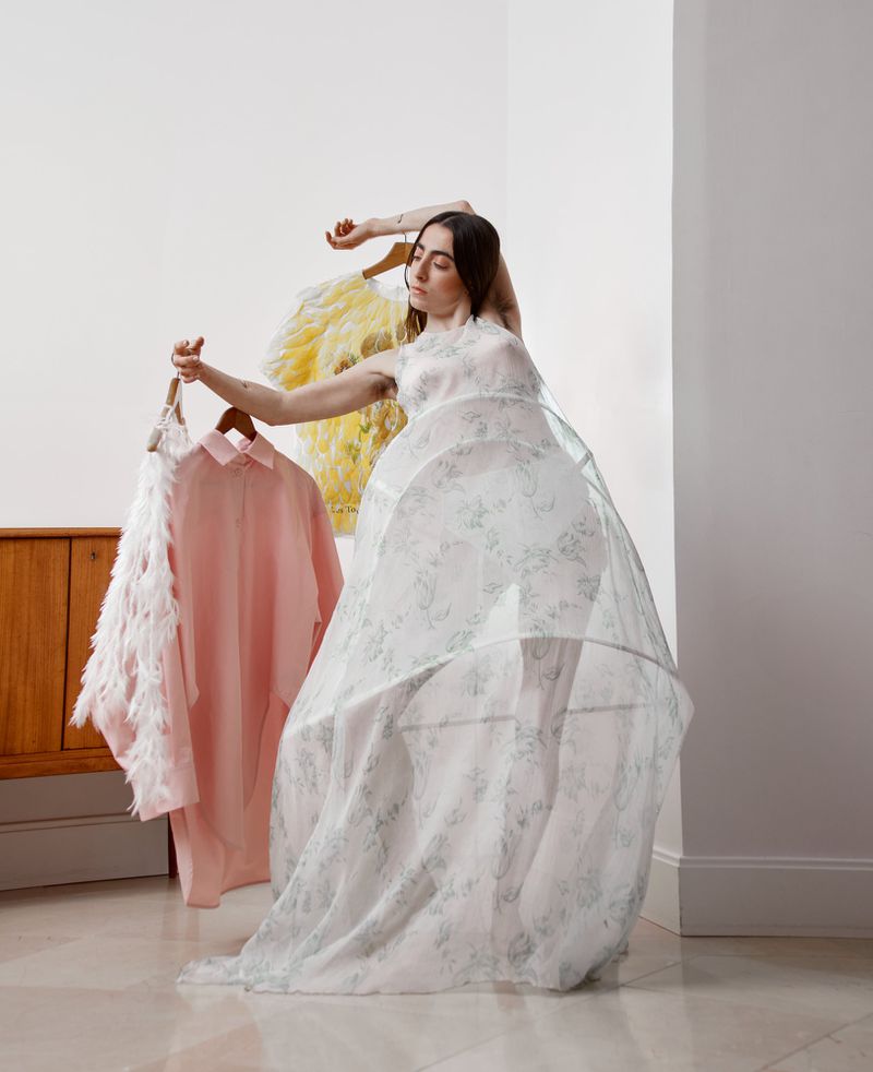 A young woman poses indoors wearing a white patterned dress with a layered tulle skirt, holding pink and white clothing on hangers.