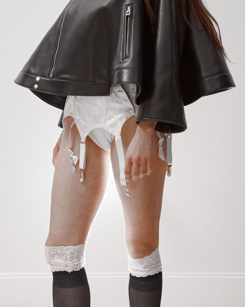 Adult wearing a black leather jacket, white garter belt, and black stockings with white lace details against a neutral background.