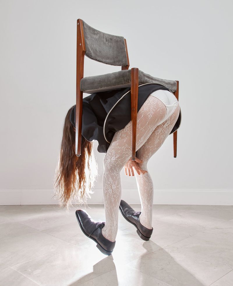 Woman bending over with chair balanced on her back. Wearing white patterned stockings, black dress and dress shoes, white backdrop.