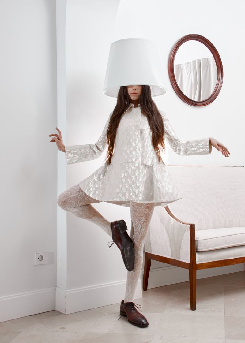 A young woman with long brown hair balances on one leg, wearing a lamp shade as a hat, a silver dress, patterned tights, and dark brown shoes.