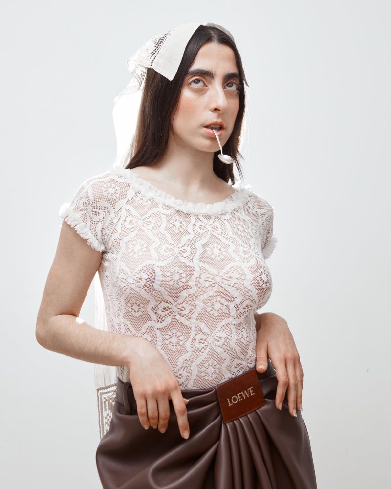 Woman posing in white lace top and brown draped skirt with the Loewe logo in a bright, neutral studio setting with a spoon in her mouth.