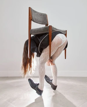 Surreal fashion image of a woman bending over wearing a chair on her back with white lace tights and black leather shoes