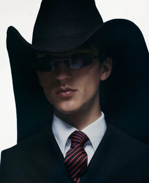Model wearing a black cowboy hat, dark sunglasses, white shirt, and striped tie against a white background for Fantastic Man.