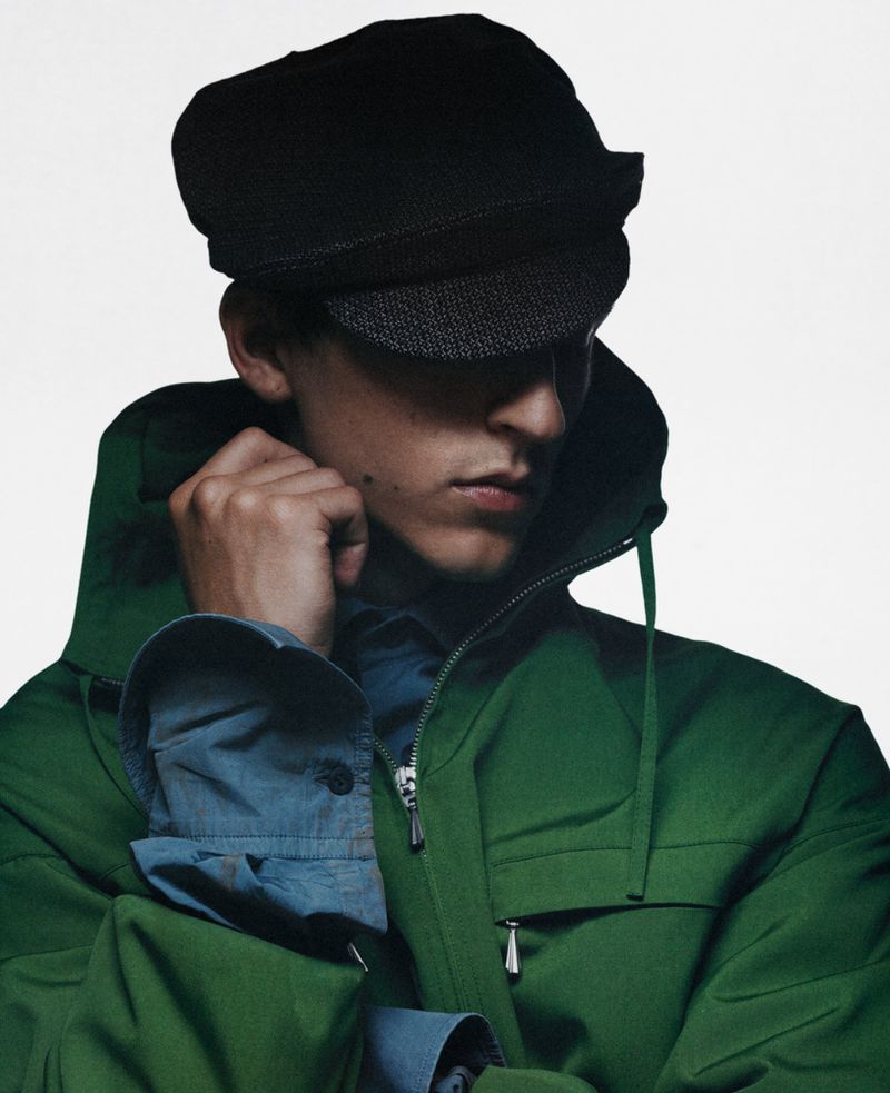Close-up portrait of a male model wearing a green hooded jacket, blue shirt, and dark flat cap against a white background.