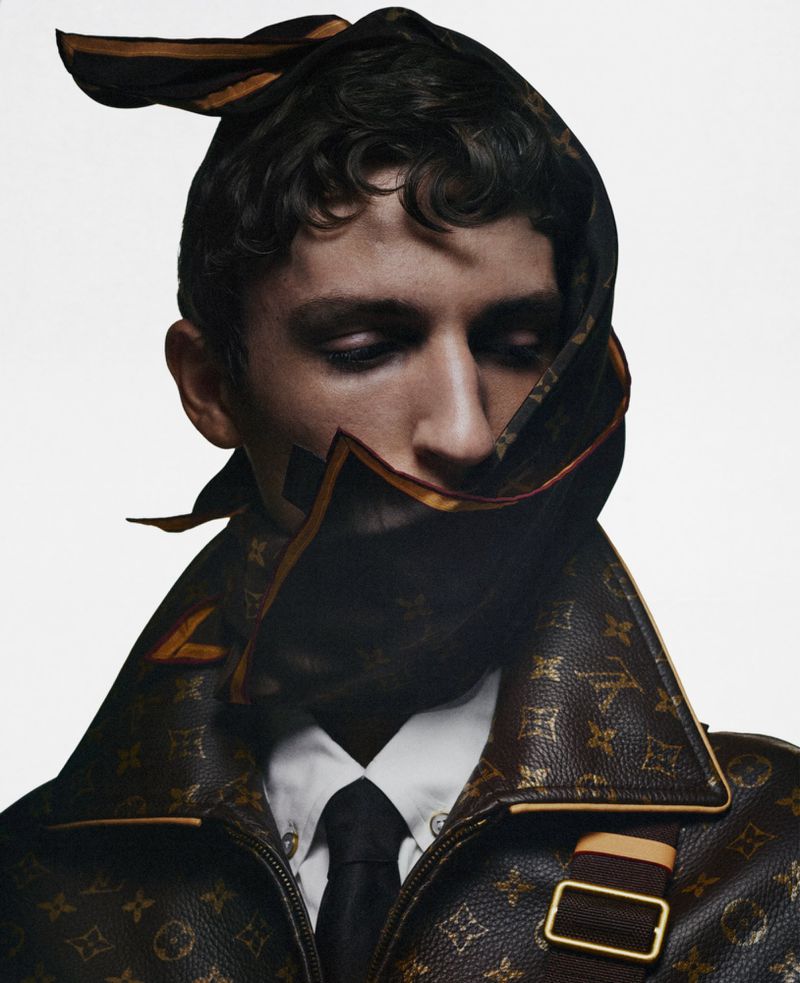 Close-up portrait of a male model wearing a monogrammed patterned jacket with a matching headscarf wrapped around his head.