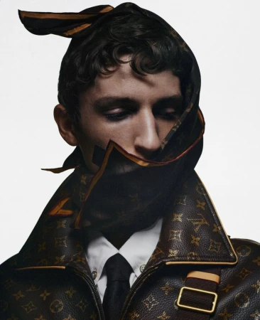 Close-up portrait of a male model wearing a monogrammed patterned jacket with a matching headscarf wrapped around his head.