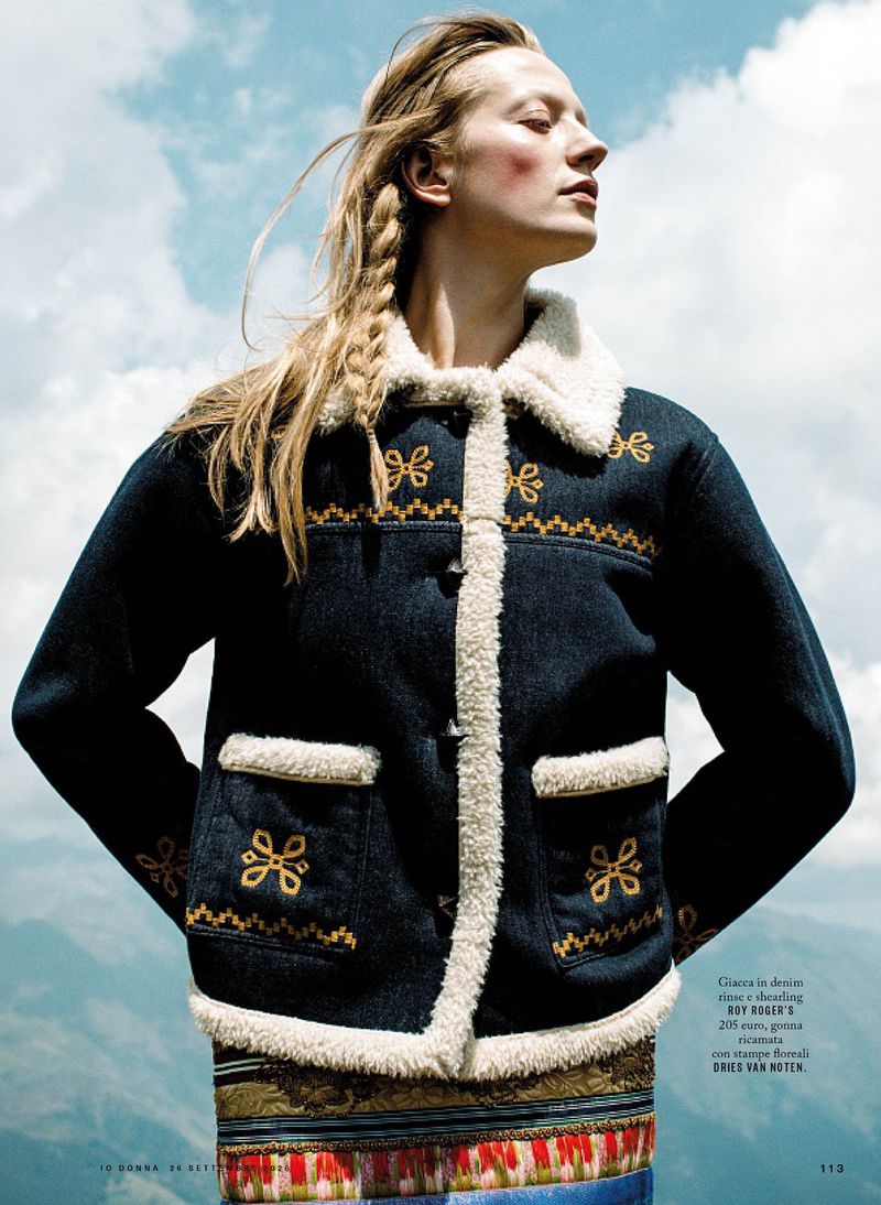 Model wearing a dark jacket with white shearling trim and yellow embroidery against an outdoor mountain and sky background.