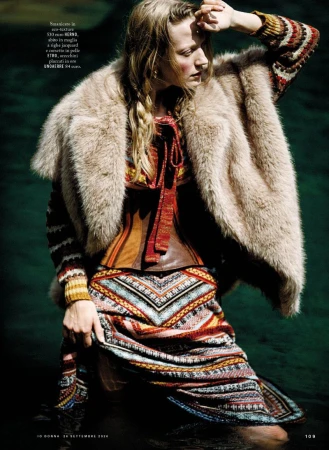 Model wearing a faux fur coat, patterned knit dress, and leather corset standing outdoors near water for iO Donna