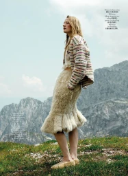 Model standing on a grassy mountain slope wearing a patterned jacket and textured skirt against a rocky peak backdrop