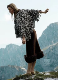 Model in a textured fringed poncho and brown skirt standing on a rocky mountain ridge overlooking distant peaks.