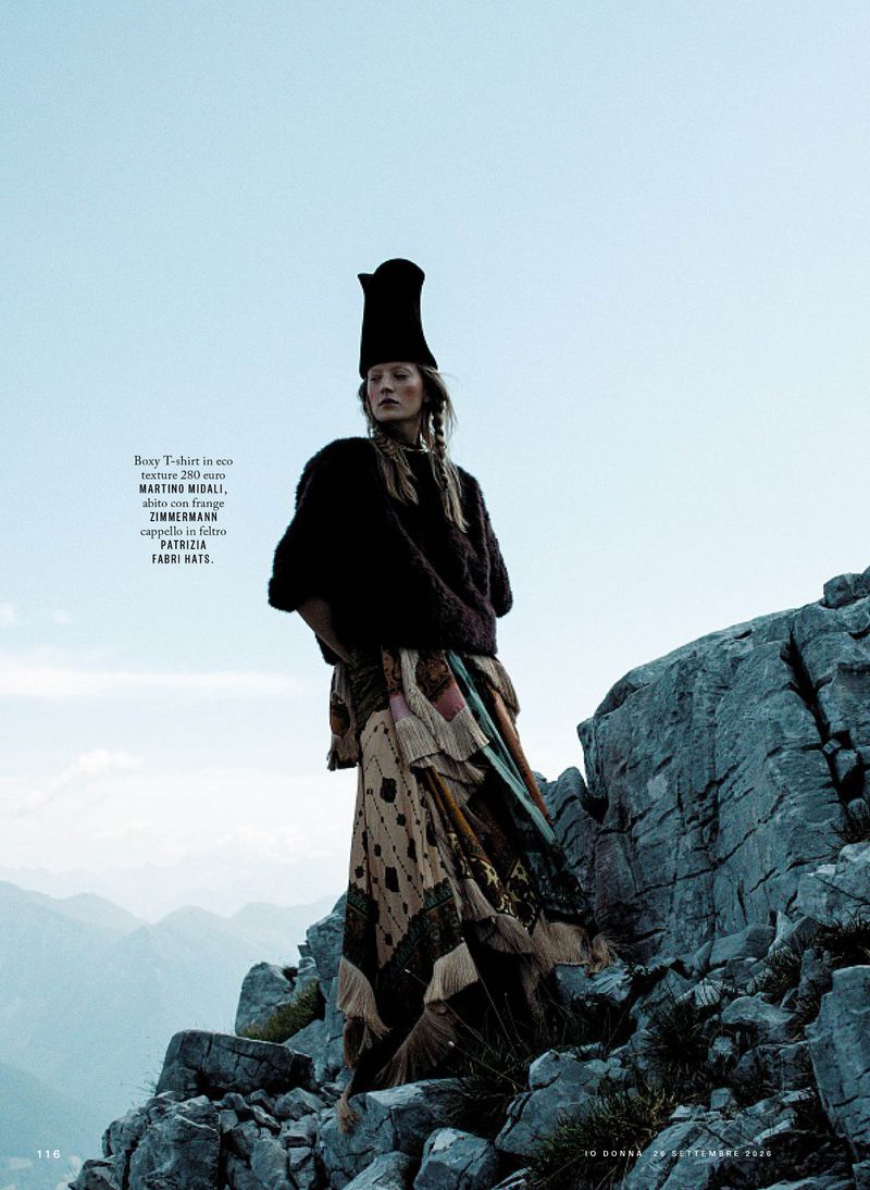 Model standing on a rocky mountain summit wearing a dark sweater, patterned skirt, and a tall black hat against a pale sky.