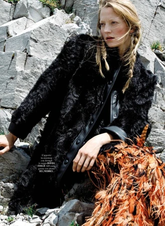 Model in a black textured coat and orange feathered skirt posed against a rocky stone cliffside.