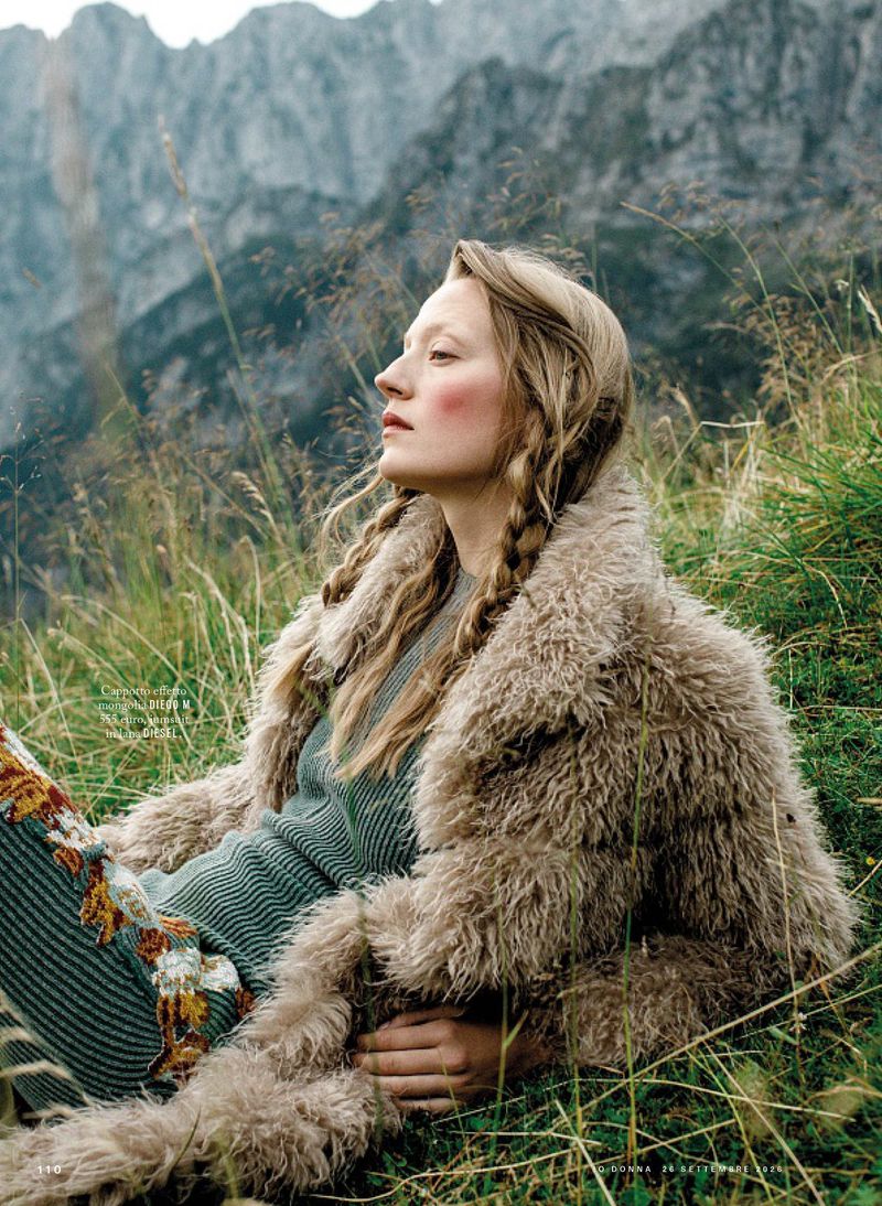 Model with braided blonde hair wearing a furry beige coat and ribbed green dress lying in a grassy alpine meadow with rocky mountains