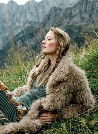 Model with braided blonde hair wearing a furry beige coat and ribbed green dress lying in a grassy alpine meadow with rocky mountains