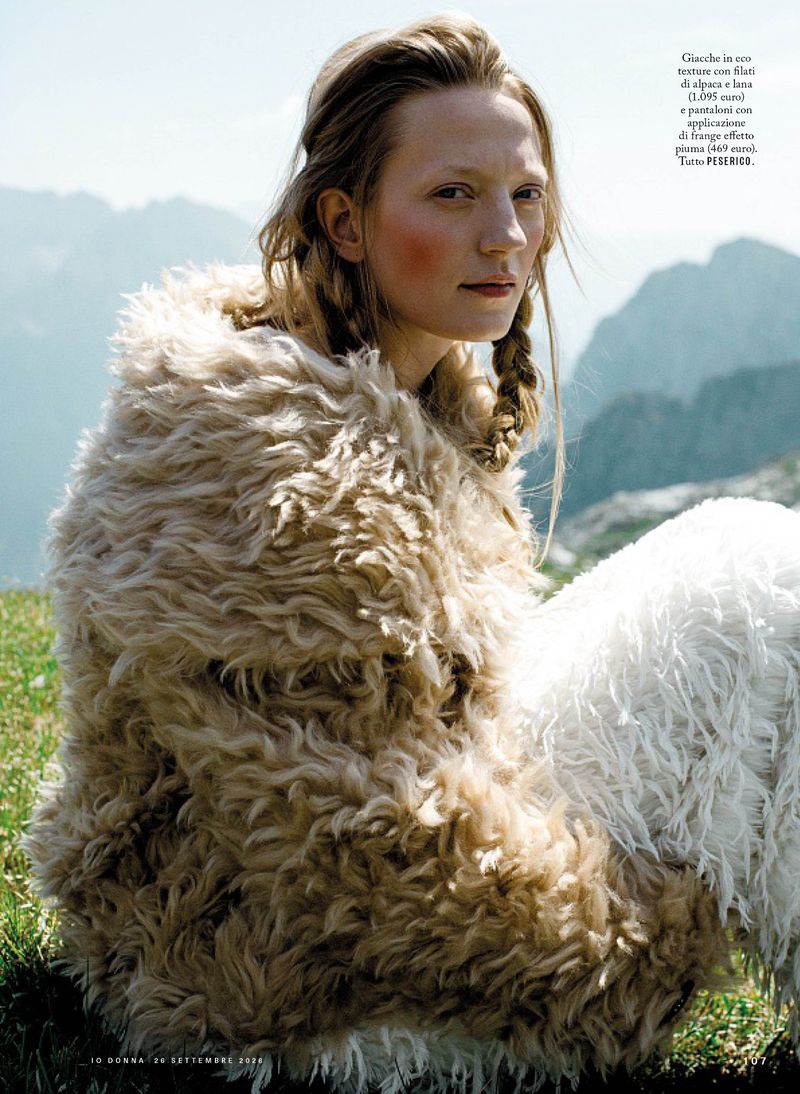 Model wearing a textured alpaca wool coat seated outdoors with mountain peaks in the background for iO Donna editorial.