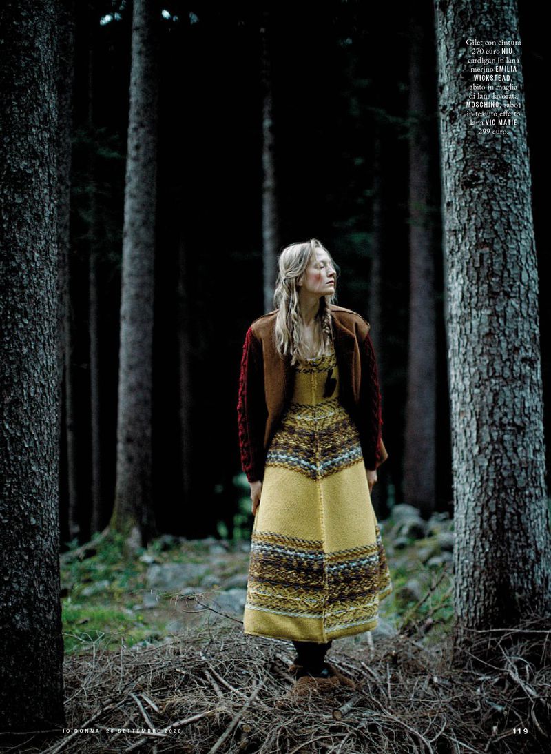 Model in yellow patterned dress and brown cardigan standing among tall pine trees in a forest setting.