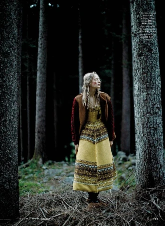 Model in yellow patterned dress and brown cardigan standing among tall pine trees in a forest setting.